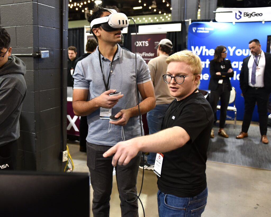 A delegate wearing a mixed-reality headset is guided by an exhibitor at a trade show stand