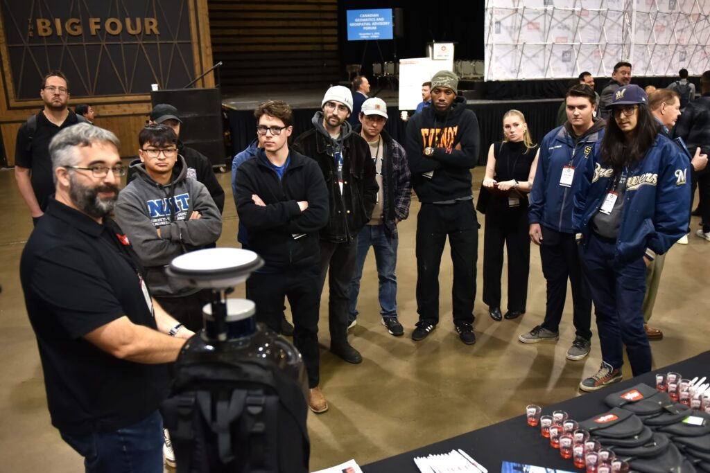 An exhibitor demonstrates a GNSS receiver on a tripod to a group of students beneath the Big Four venue lettering